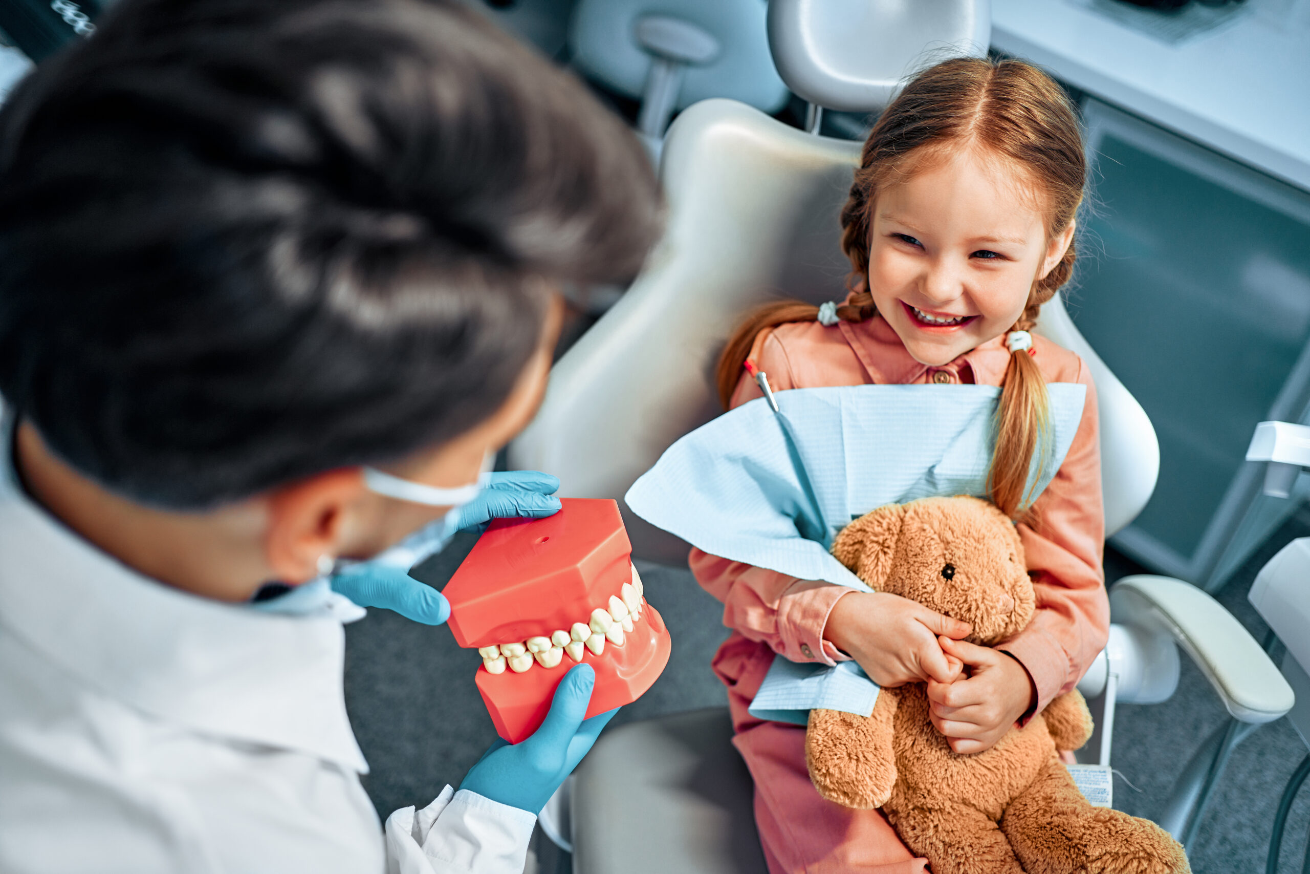 Young girl in a dental chair smiling at a dentist who holds a large teeth model and a red tray, with a plush teddy bear in her lap.